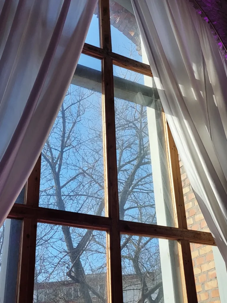 Detal dużego okna z białymi zasłonami w historycznym budynku, z widokiem na warszawską zieleń i architekturę miejską – tło dla motta BMB Realty